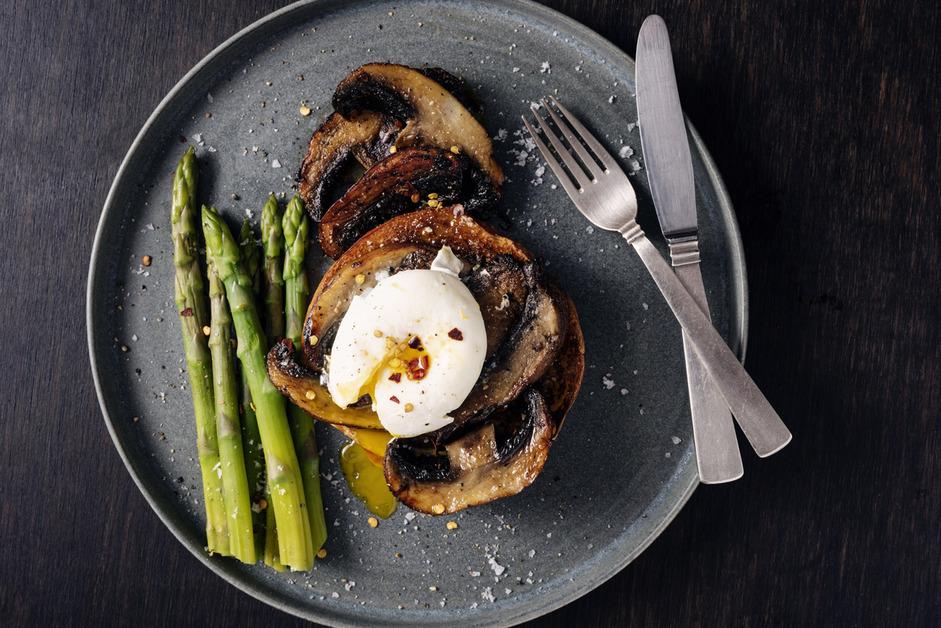 A gray plate of asparagus, cooked portobello mushroom slices, and eggs with a fork and a knife on a black table. 