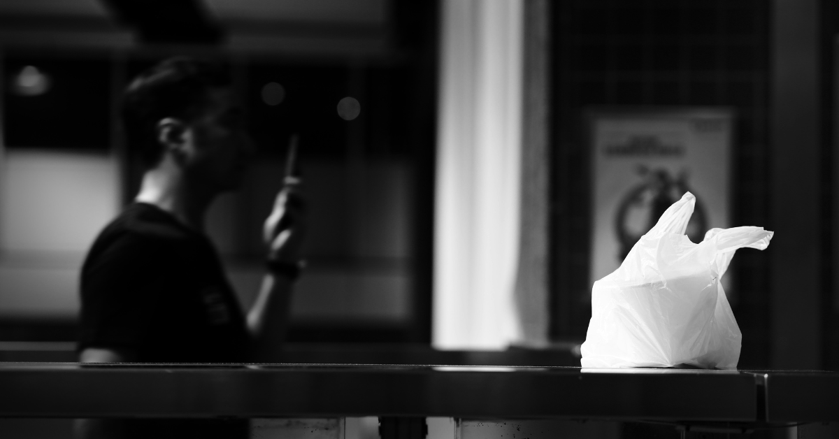 A black and white image features a man waiting in the background while his takeout items are placed into a white plastic bag and sat on a counter