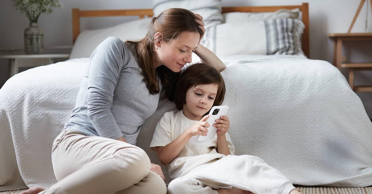 A mother sitting on the bedroom floor with her daughter relaxing and looking at a smart phone together