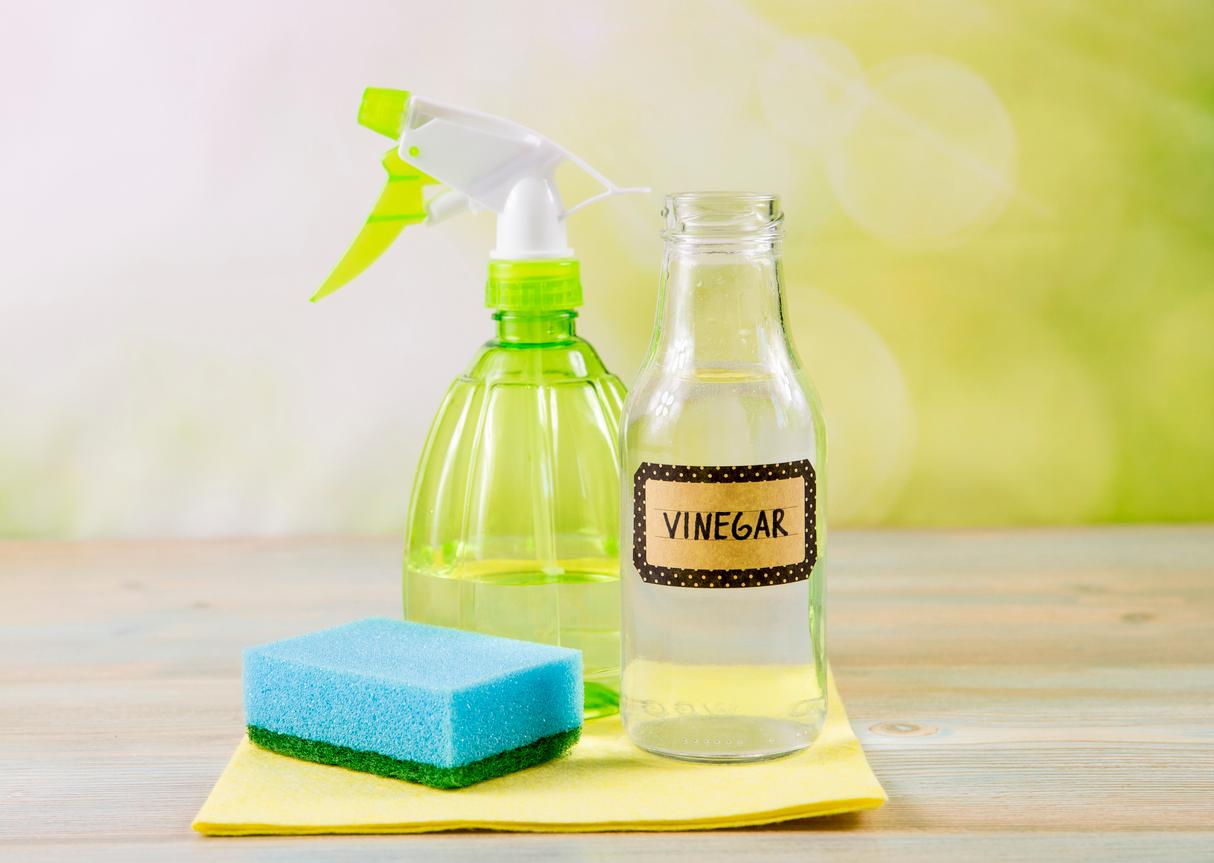 A bottle of vinegar is pictured beside a green spray bottle and a blue sponge atop a wooden table.