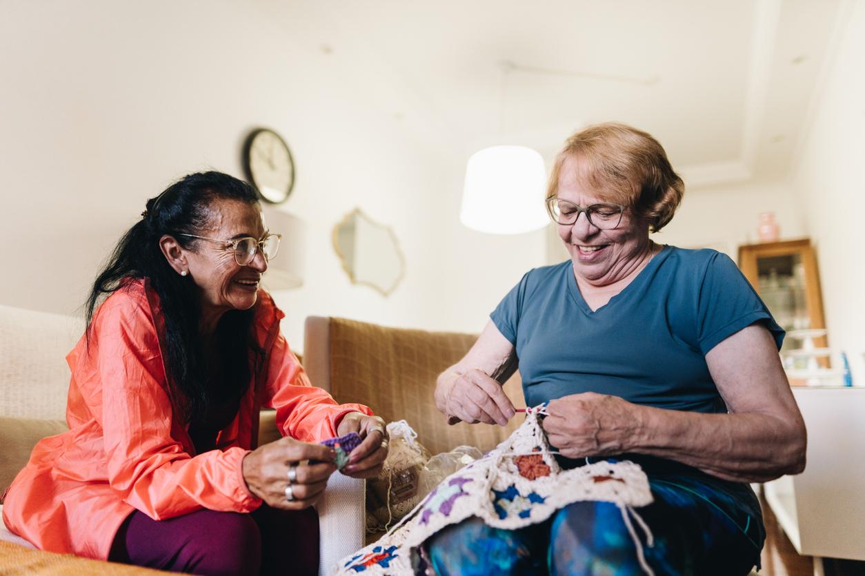 Older adults sit in a living room and smile as they crochet together.