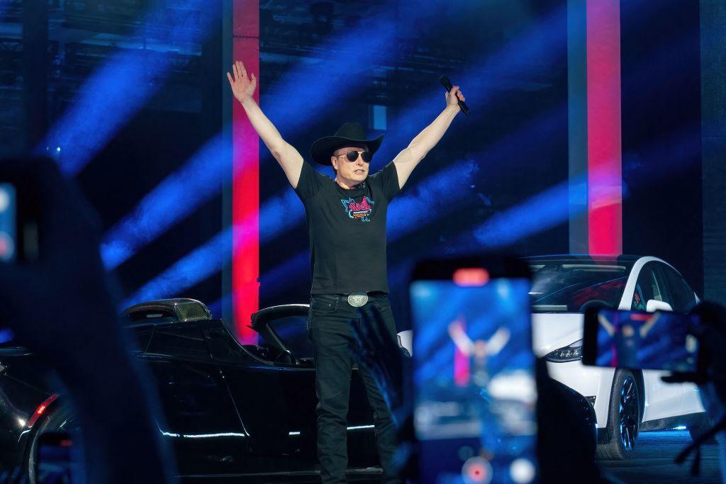 Tesla CEO Elon Musk, wearing an all-black outfit and black cowboy hat, throws his arms in the air in front of a crowd at the Tesla Giga Texas manufacturing "Cyber Rodeo" grand opening in April 2022 in Austin, Texas.