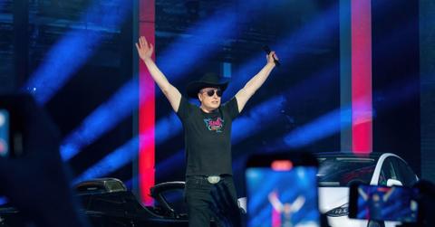 Tesla CEO Elon Musk, wearing an all-black outfit and black cowboy hat, throws his arms in the air in front of a crowd at the Tesla Giga Texas manufacturing "Cyber Rodeo" grand opening in April 2022 in Austin, Texas.
