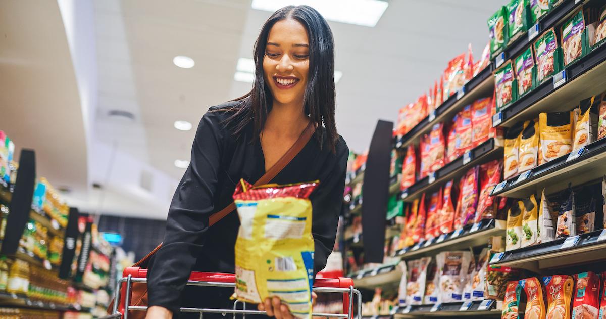 Smiling woman adds bag of chips to her cart in a grocery store aisle.