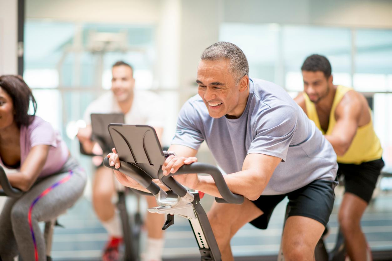 A smiling older vegan athlete rides an exercise bike in a fitness class with other smiling adults.