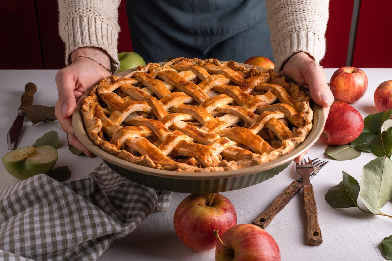 An apple pie is pictured above a table with loose apples, green leaves, a napkin, and forks.