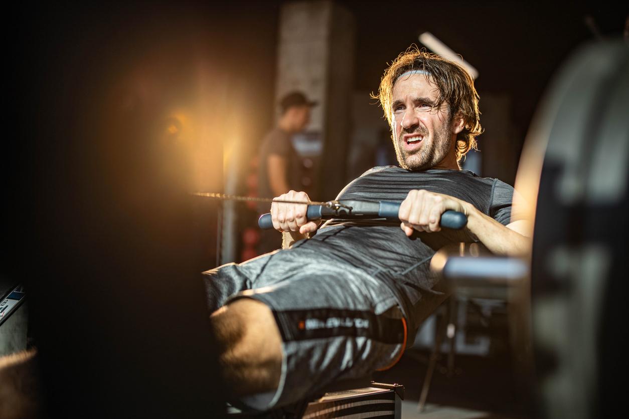 A man lays all the way back while rowing on an ergometer inside a gym.