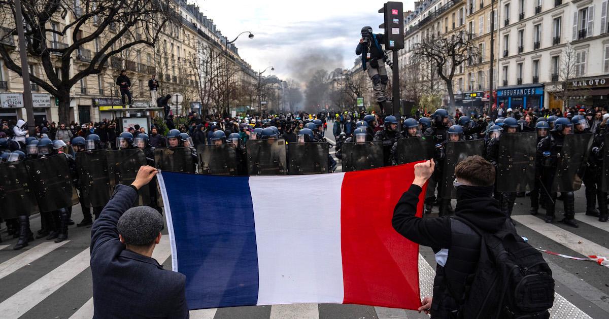 On a street in France, two protesters hold up a French flag in an intersection, facing several dozen police officers wearing helmets and shields. 