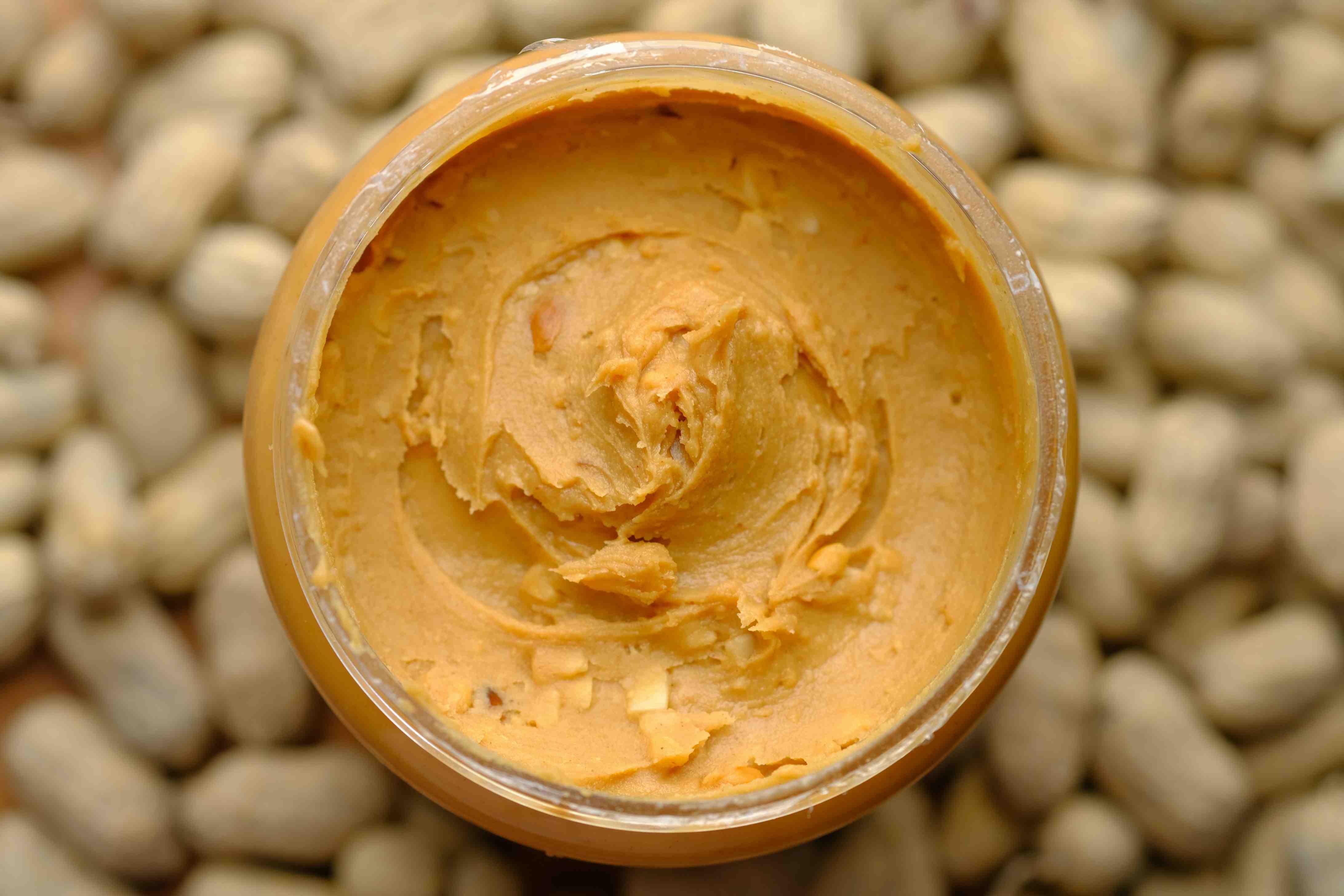 A jar of peanut butter appears full to the top, above a blurry pile of peanuts.