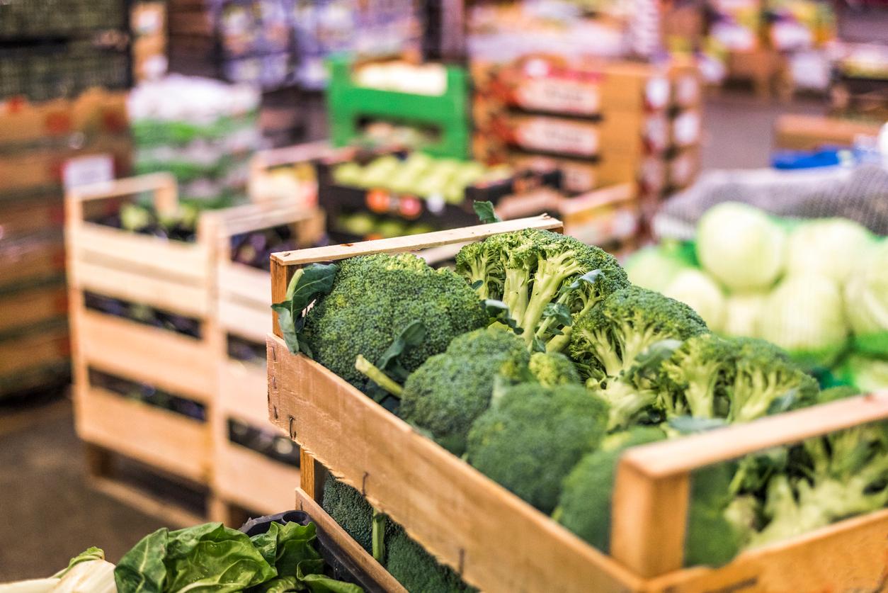 Heads of broccoli are pictured in a wooden crate with other vegetables blurred in the background of a market.