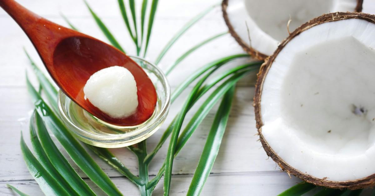 A wooden spoon scoops a bit of coconut oil from a jar next to a split coconut