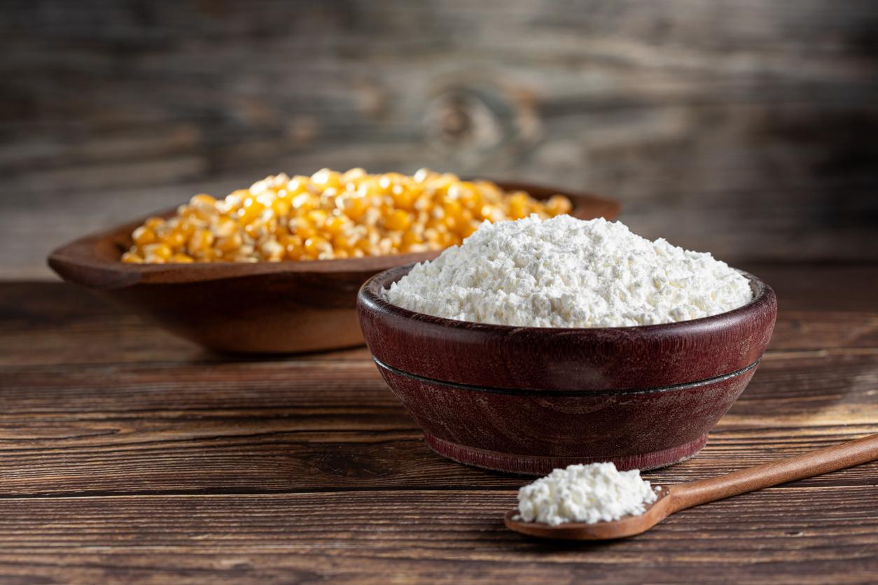 A wooden bowl of cornstarch is pictured beside a wooden spoonful of cornstarch and next to a wooden bowl of corn.