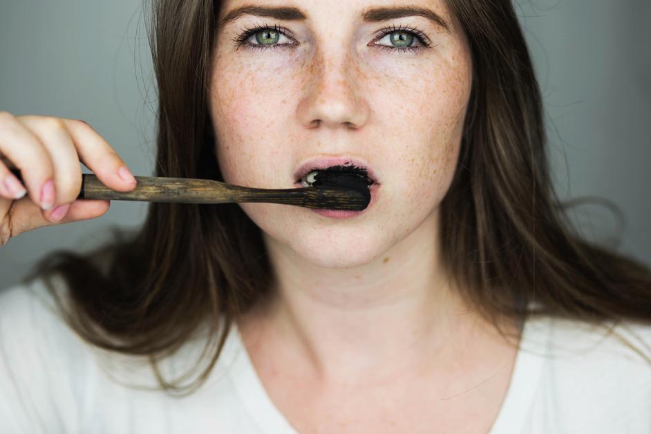 A woman with brown hair, freckles, and green eyes brushes her teeth with charcoal toothpaste in front of a gray background. 