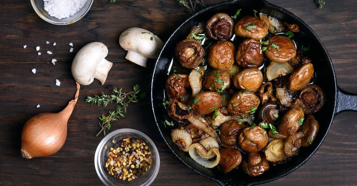 Skillet full of delicious looking sautéed mushrooms with a variety of ingredients scattered on a wooden table top. 