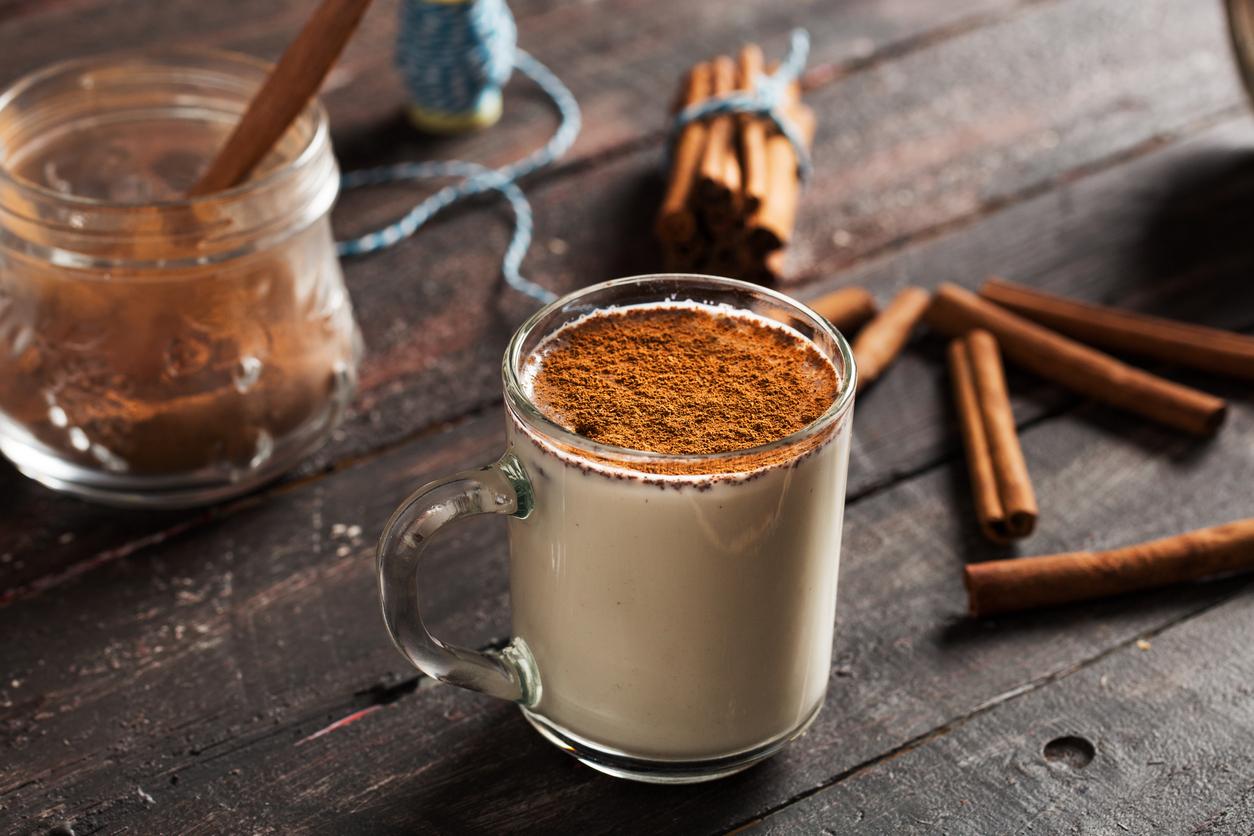 A cup of coffee with cinnamon sprinkled on top beside cinnamon sticks laying on a table.