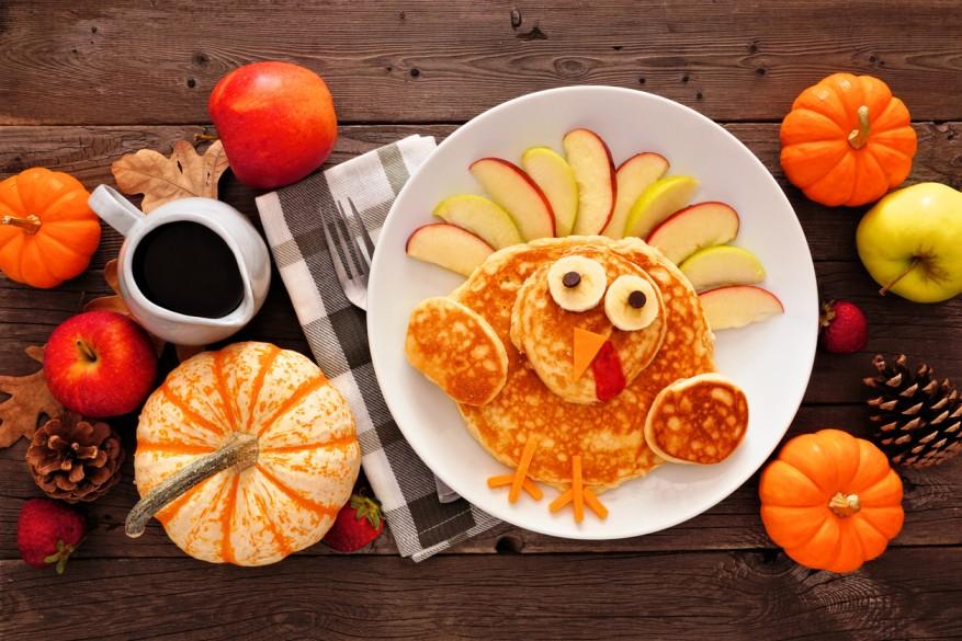 Pancakes with apple slices above them are pictured on a plate beside pumpkins of various sizes.