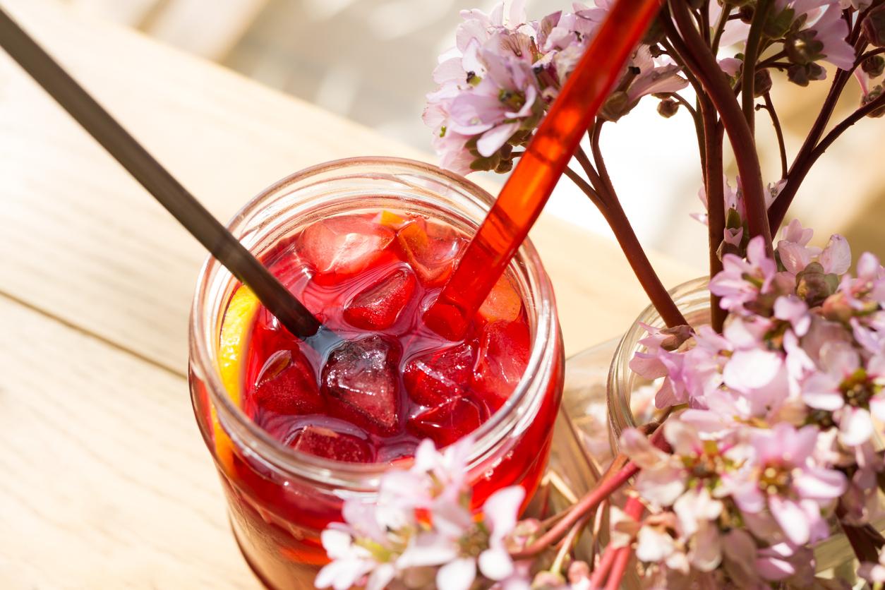 Red hibiscus tea is prepared in a glass jar beside flowers.