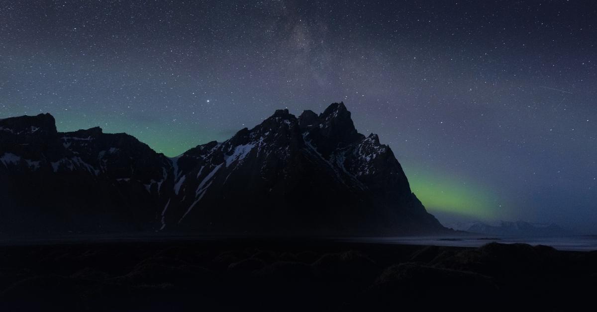 Photograph of the Northern Lights behind a mountain on a black beach in Iceland. 