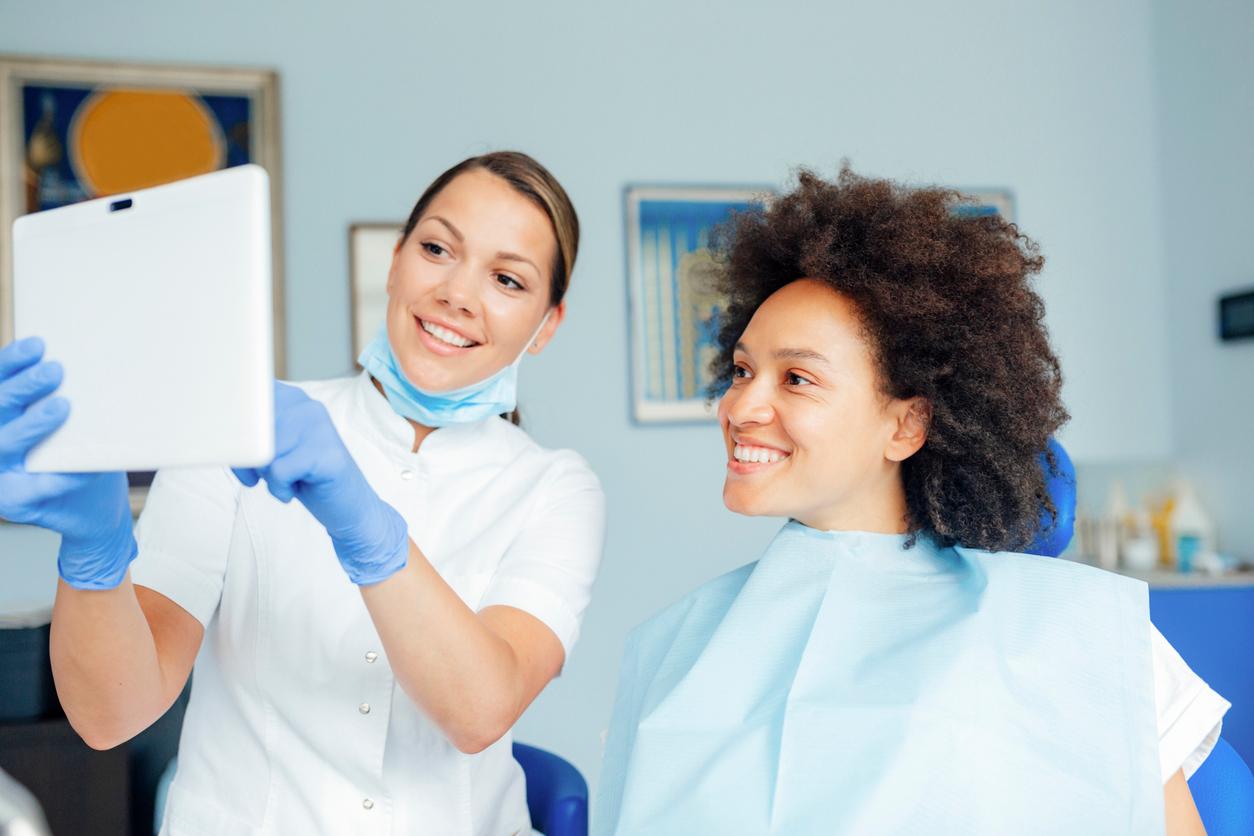 A smiling patient with a blue bib looks at a tablet held by a smiling dental hygienist.