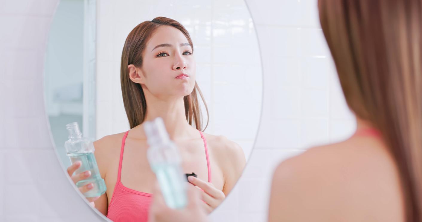 A woman holds a mouthwash bottle in one hand while viewing herself in the mirror as she swishes mouthwash in her mouth.