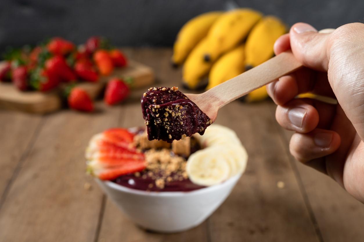 A person holds a wooden spoonful from an açaí bowl atop a wooden table with strawberries and lemons in the background.