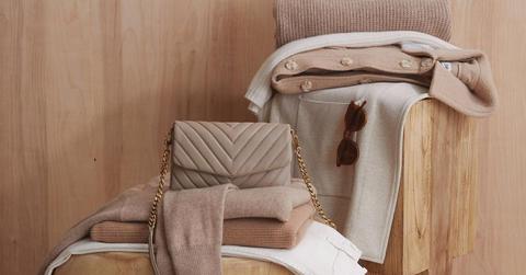 Quince product photo of stacked beige and tan sweaters, a purse, and a pair of sunglasses in front of a wooden background