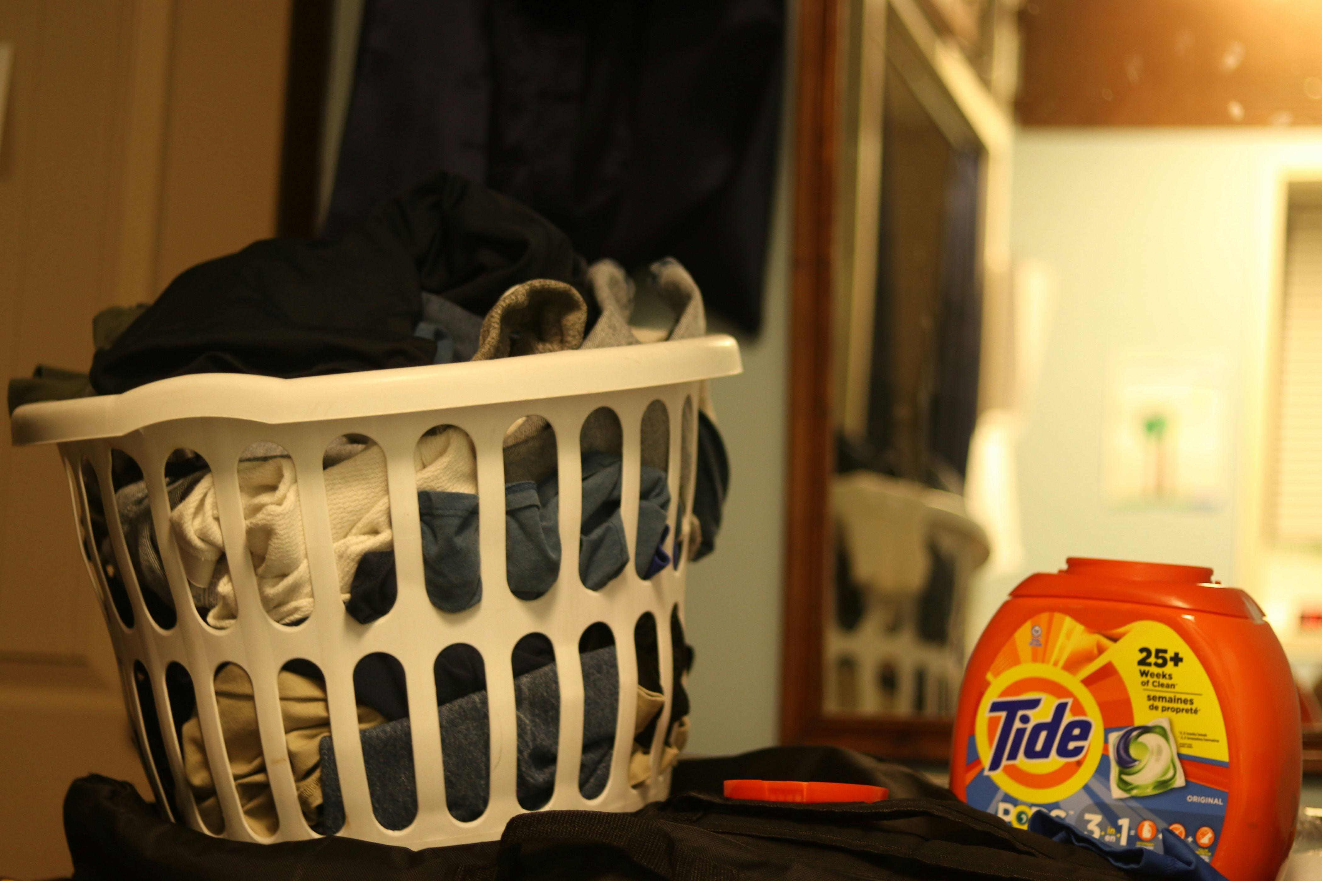 A white laundry basket with holes in it is pictured filled with laundry beside a package of Tide Pods laundry detergent.