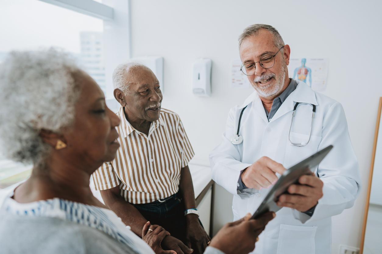 A smiling doctor talks to two patients while pointing to information on the doctor's tablet.