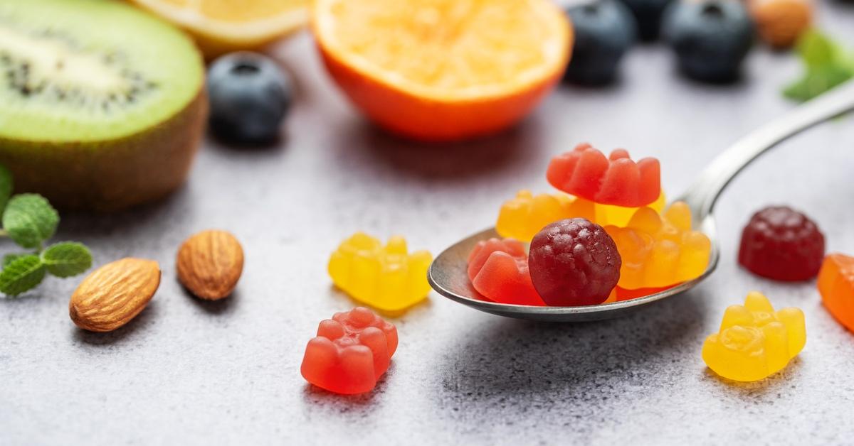 Clusters of multicolored gummies in a spoon next to almonds, kiwi, and orange on a table.