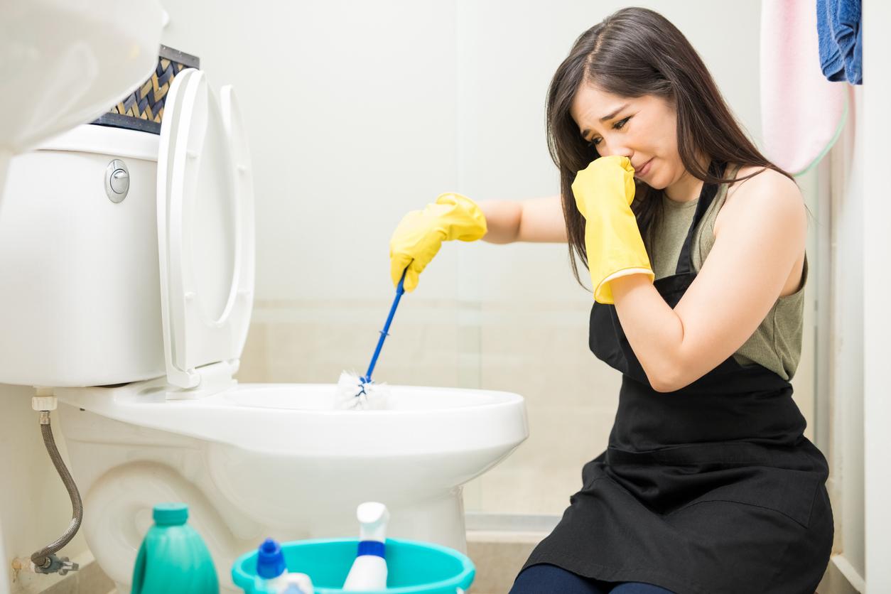 A woman wears yellow rubber gloves while brushing a toilet bowl clean and holding her nose with her other hand.