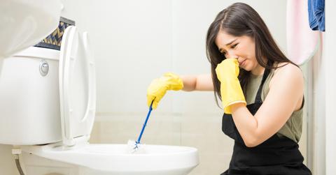 A woman wears yellow rubber gloves while brushing a toilet bowl clean and holding her nose with her other hand.