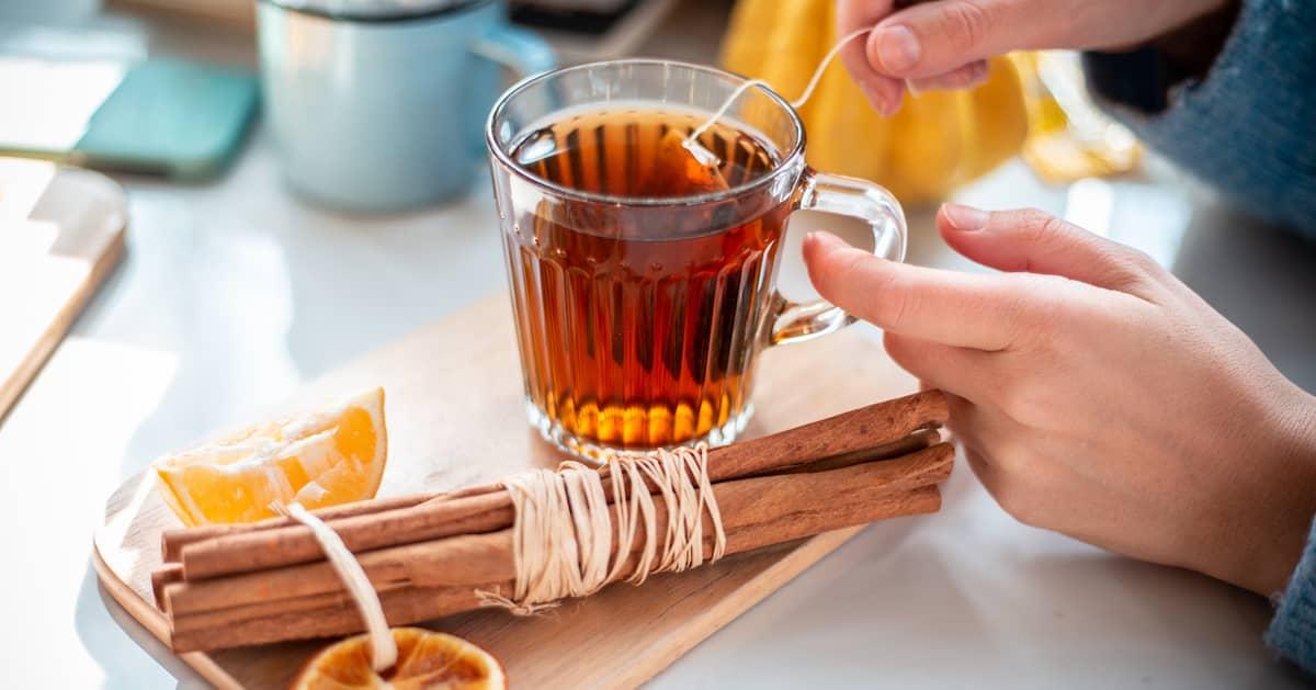 Closeup of a person brewing a cup of cinnamon-infused tea, with cinnamon sticks and slices of lemon next to the cup