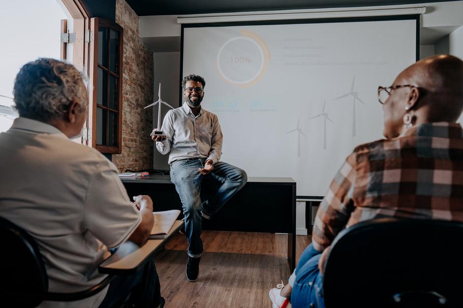 A man sits on a desk while presenting a project on wind turbines to a room of people. 