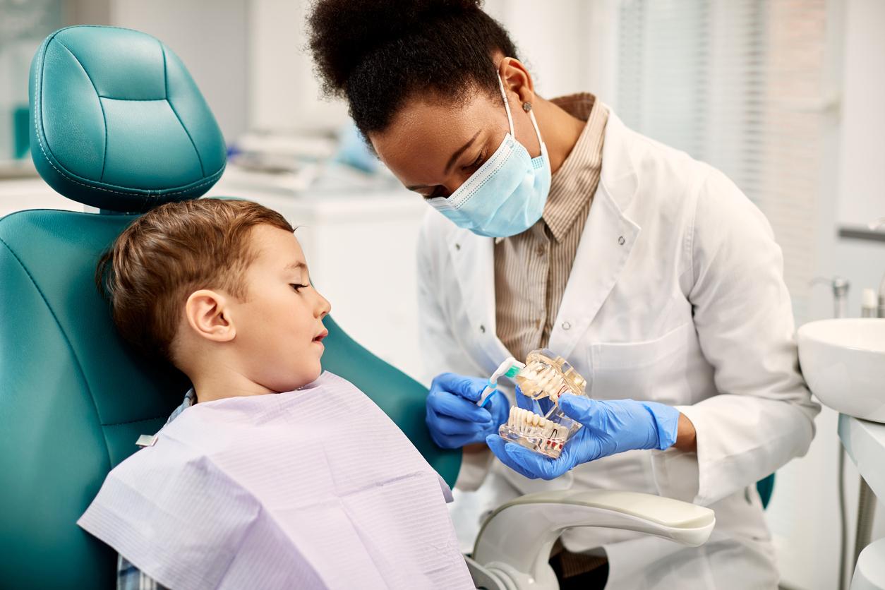 A female dentist explains healthy brushing techniques to a young boy using a mouth model.