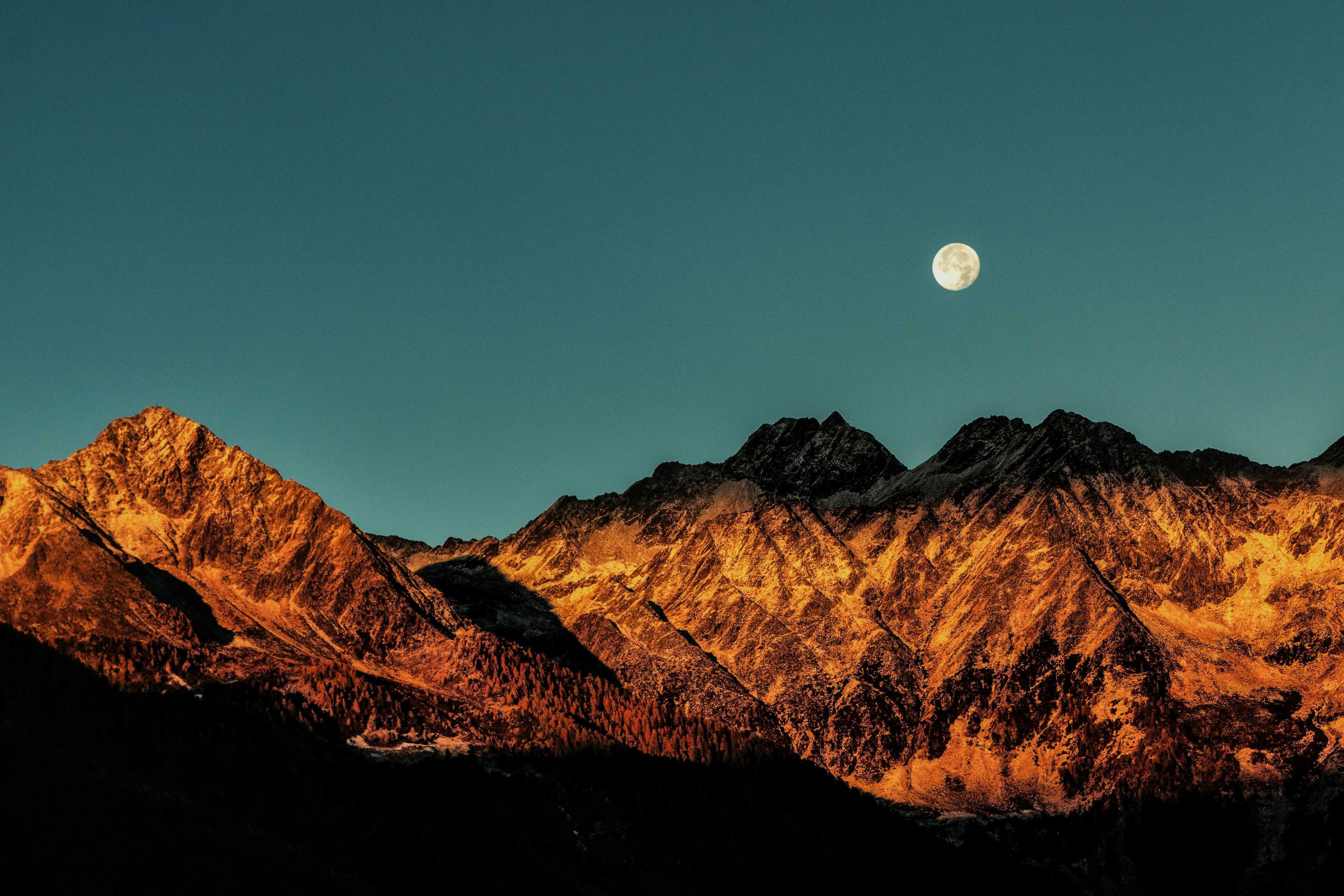 A moon appears over mountains in front of a turquoise sky at sunrise. 