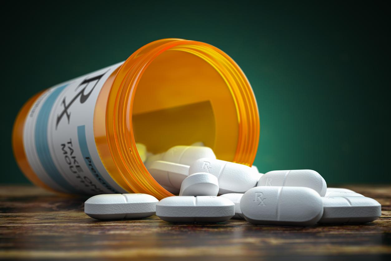 A pill bottle spills white pills with "RX" labeled on each pill on top of a wooden table beside a green wall.