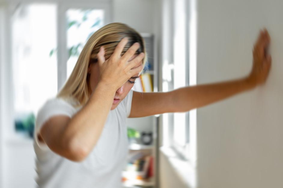 Blonde woman in white shirt presses her hand against the wall while grasping her forehead in pain. 