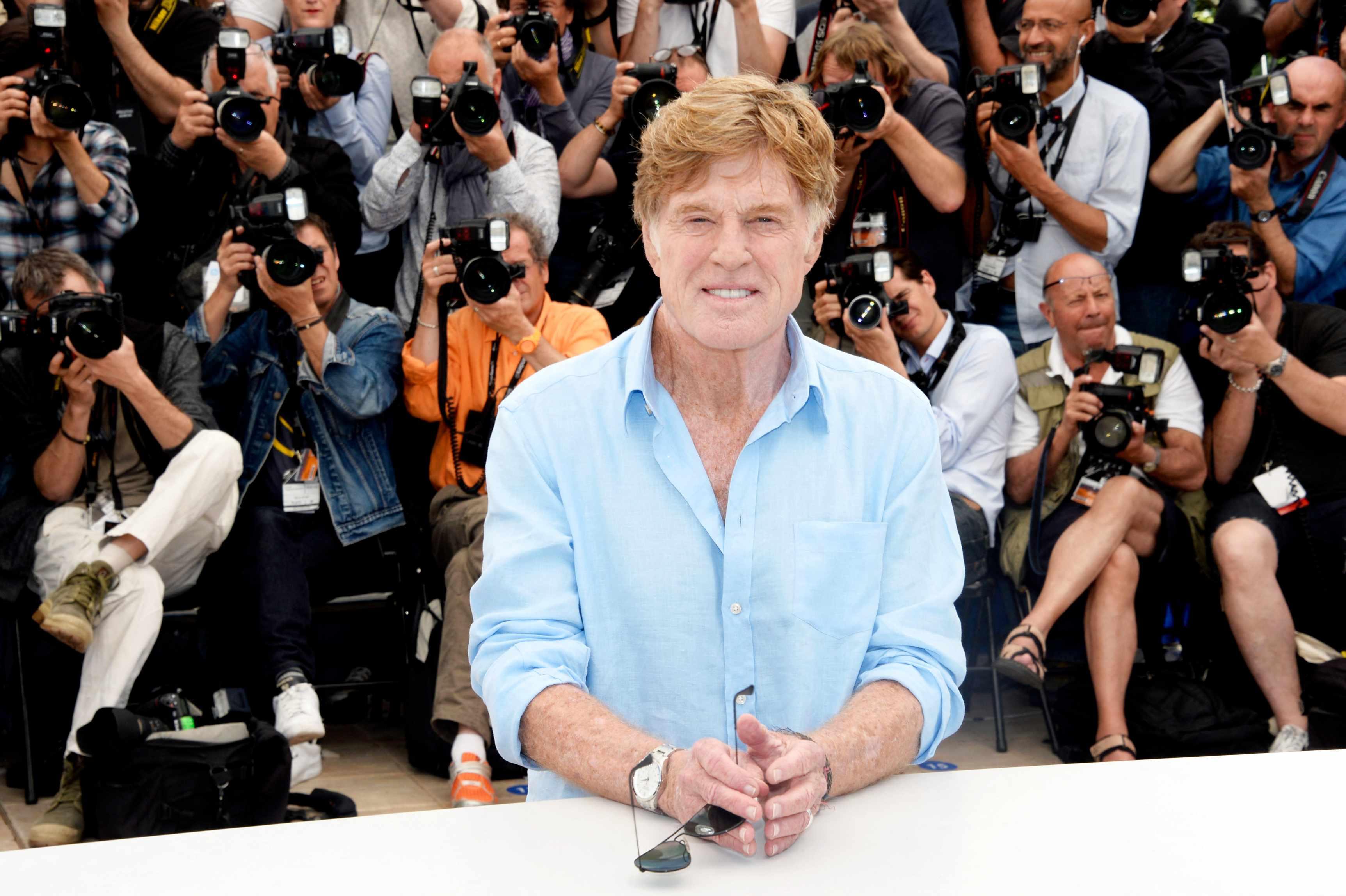 Robert Redford is pictured in a blue shirt standing in front of media members holding cameras.