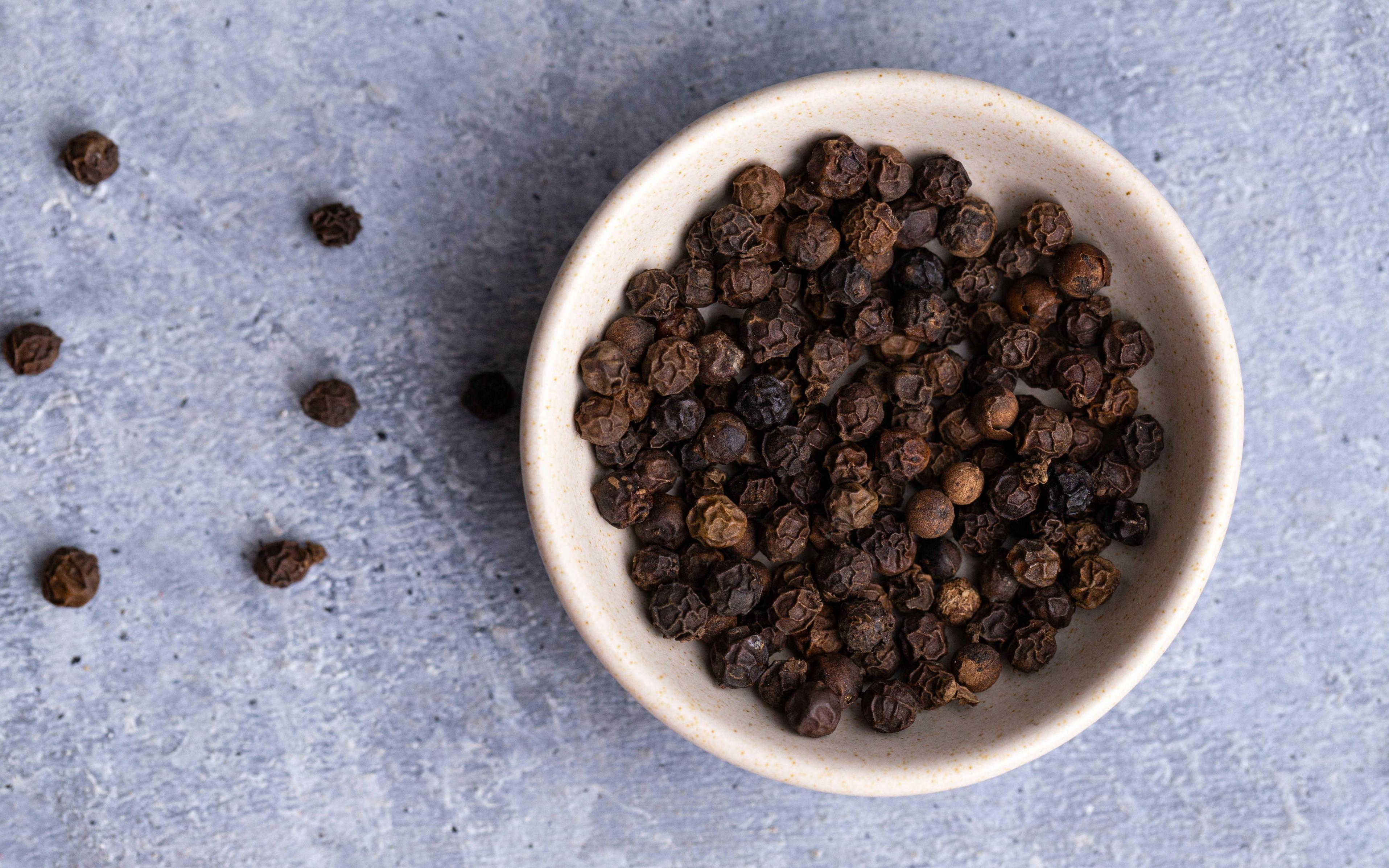 Black peppercorns are pictured in a white ceramic bowl, with some peppercorns loose on the table beside the bowl.