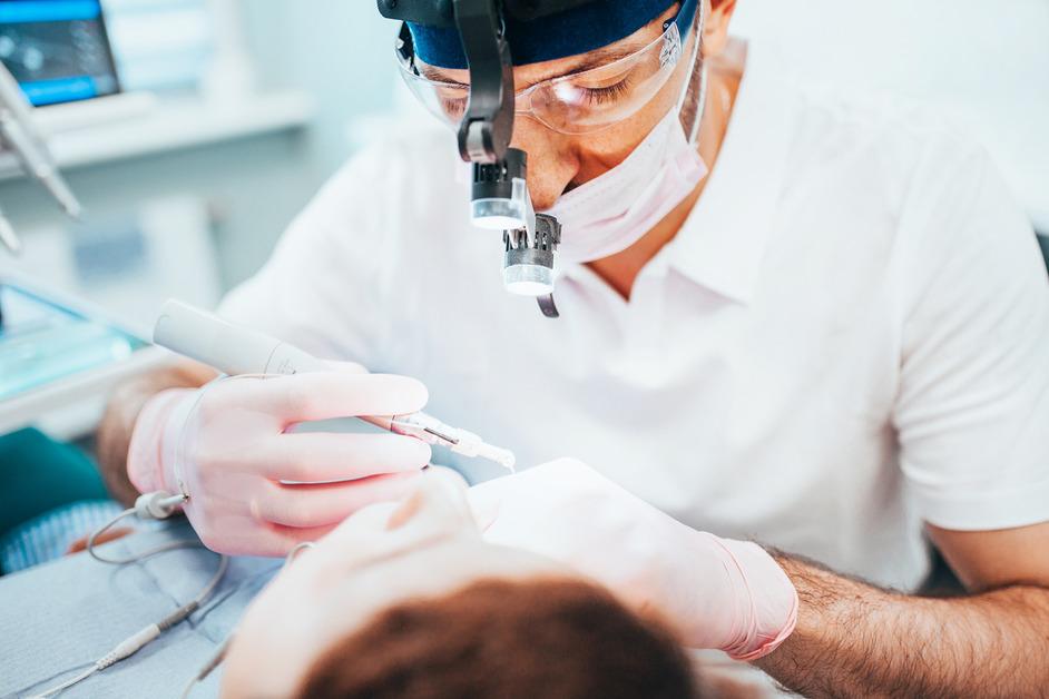 A dentist wearing a mask and a light on his head uses a tool while a patient sits in the chair. 