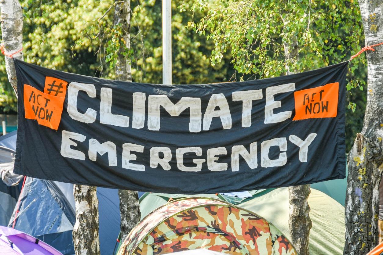 A handmade banner that reads "Climate Emergency, #actnow, act now" is hung between two trees