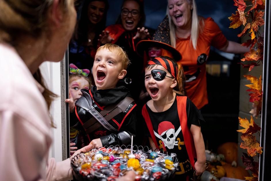 An over-the-shoulder view of a woman holding a bowl of candy while two kids in pirate costumes smile at the door with more people behind them. 