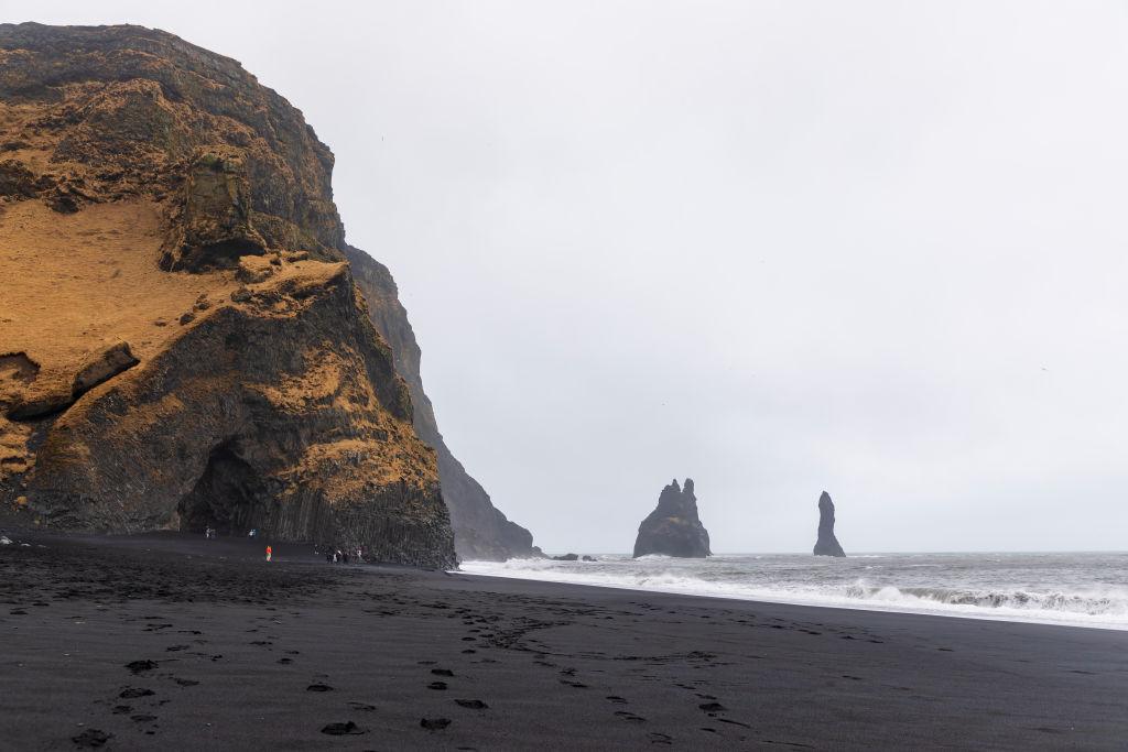 Reynisfjara Beach as seen in April 2023 with its trademark black sand, a large hill and cave to the left of the photo, and waves crashing to the shore on the right side of the photo. 