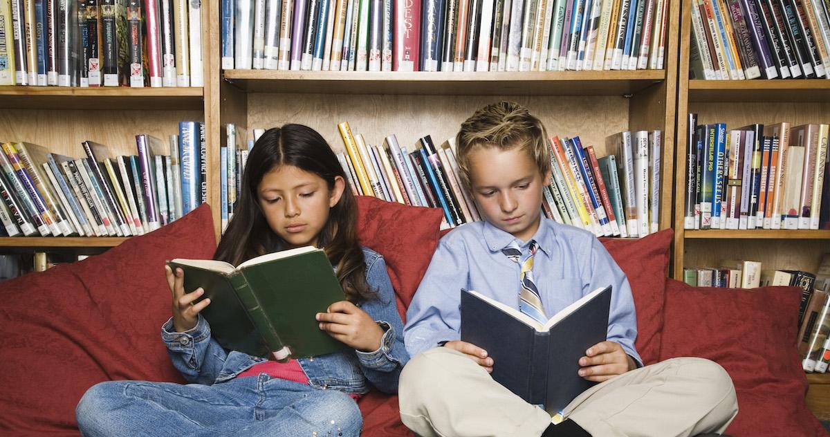 Two children reading books in a library in front of a shelf.