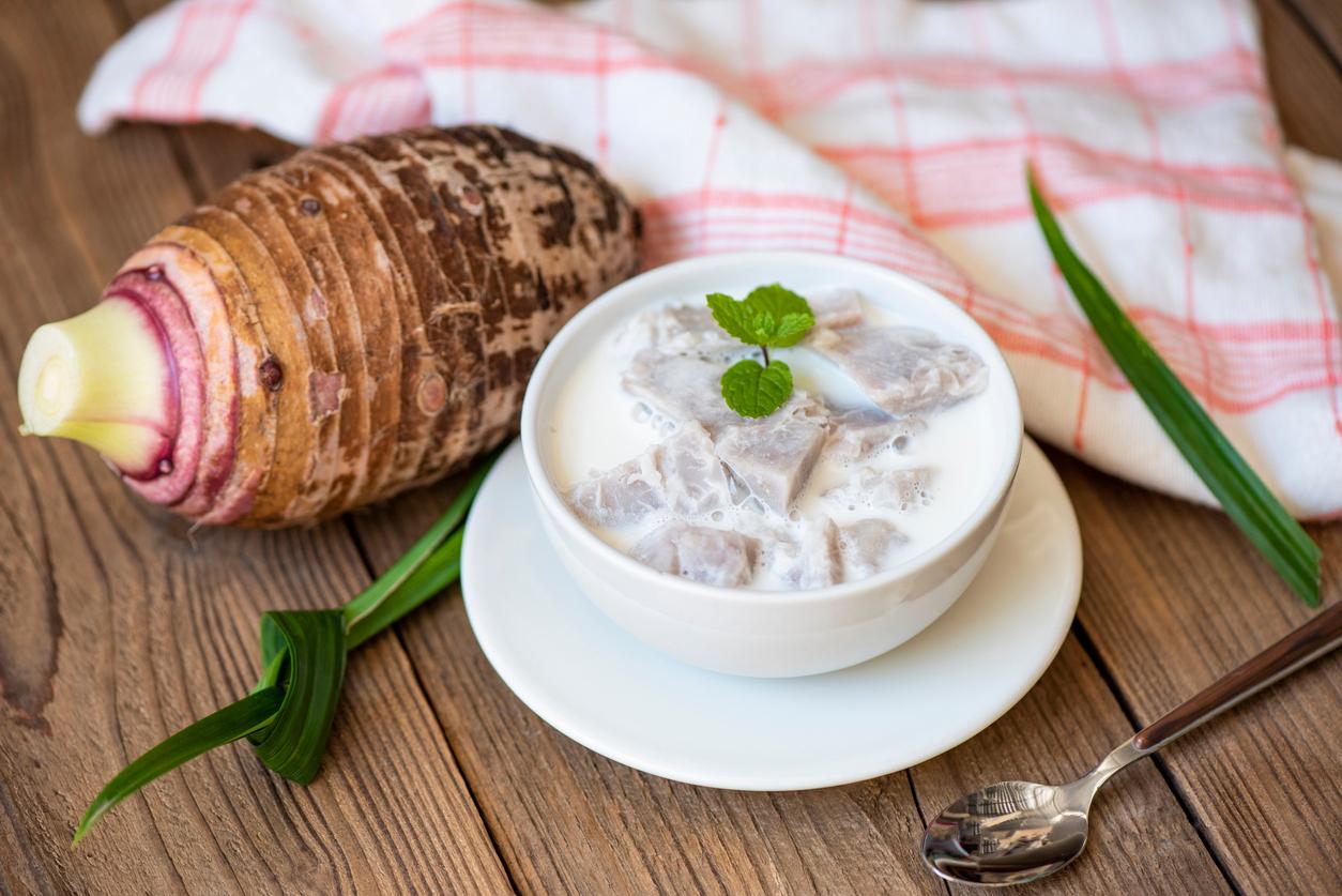 A taro root is placed on a table beside a taro-based dessert in front of a pink-and-white cloth.