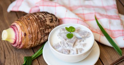 A taro root is placed on a table beside a taro-based dessert in front of a pink-and-white cloth.