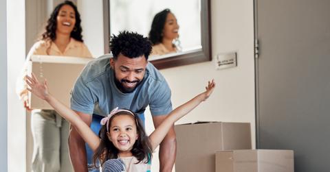 A smiling family enters a new home while a young child spreads her arms inside of a box.