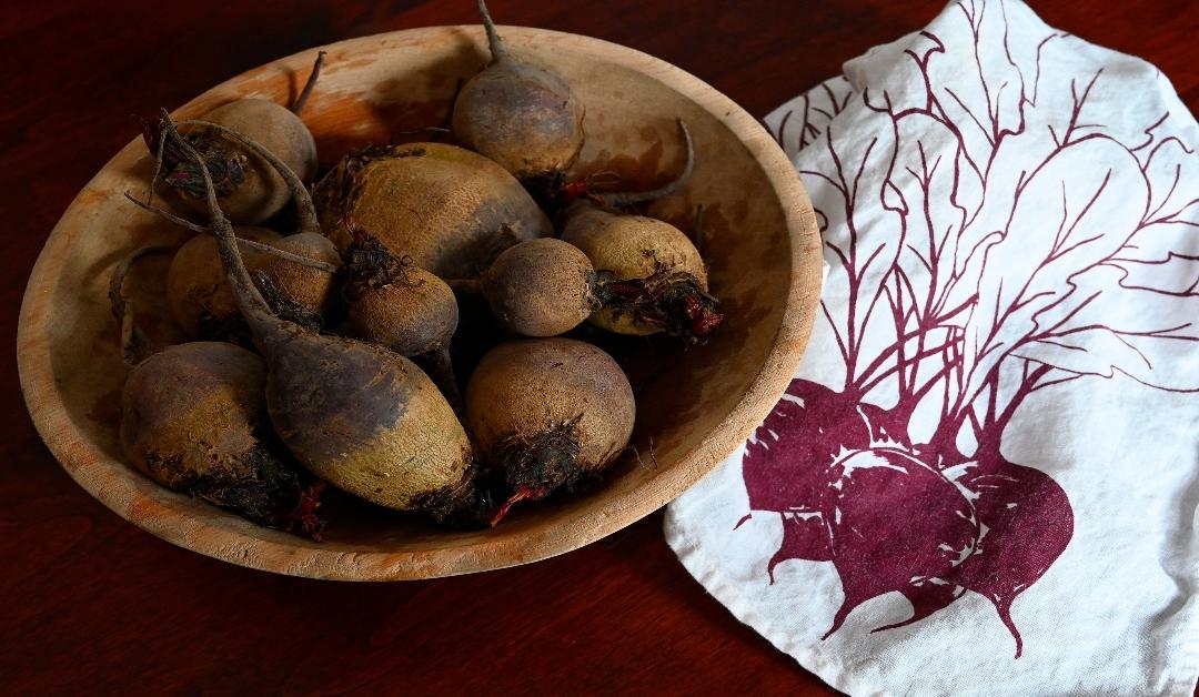 A wooden bowl of beets sitting on a table from Rosemont Grocery store with a beet napkin made by Madder Root Maine