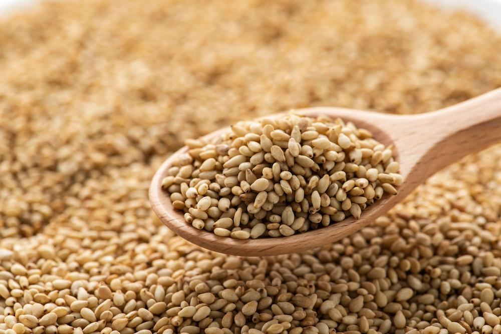 A wooden spoon full sesame seeds over a pile of the seeds. 