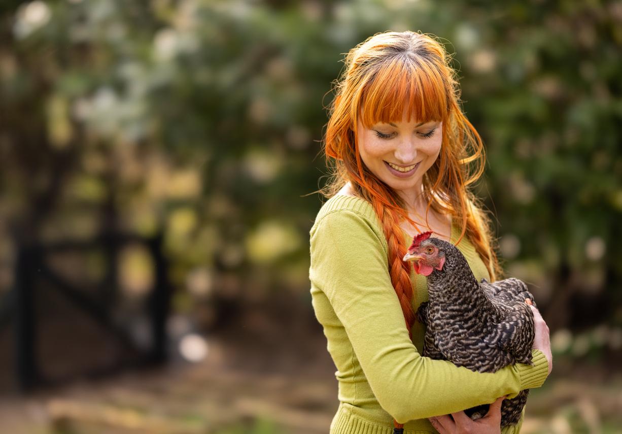 A smiling vegan woman looks down upon a chicken, which she is holding in her arms.